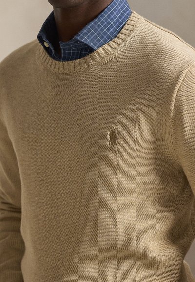 Beige strikkegenser med ribbet rund hals, med en liten brun logoemblem. Lagvis over en blå rutete skjorte.