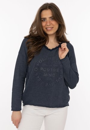 Lächelnde Frau mit langen braunen Haaren, die einen dunkelblauen Pullover mit dem Schriftzug „Positive Mind Good Vibes“ trägt, kombiniert mit weißen Hosen, vor weißem Hintergrund.