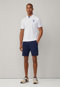 Biele polo tričko s modrým golierom a logom, skombinované s námorníckymi šortkami s potlačou. Doplnené béžovými a čiernymi športovými topánkami.