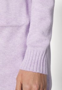 Lavendelfärgad stickad tröja med ärm och ribbat manschett, som visar en mjuk textur och slät yta, delvis täckande en hand.