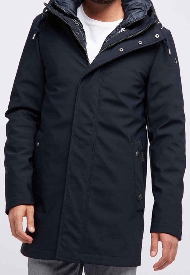 Homme portant un parka zippé bleu marine foncé avec capuche, debout les bras détendus le long du corps, portant une chemise blanche et un pantalon gris.