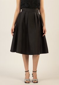 Jupe noire de longueur genou avec des plis, fabriquée en un matériau lisse. Présente une taille ajustée et une silhouette fluide. Associée à des talons noirs à lanières.