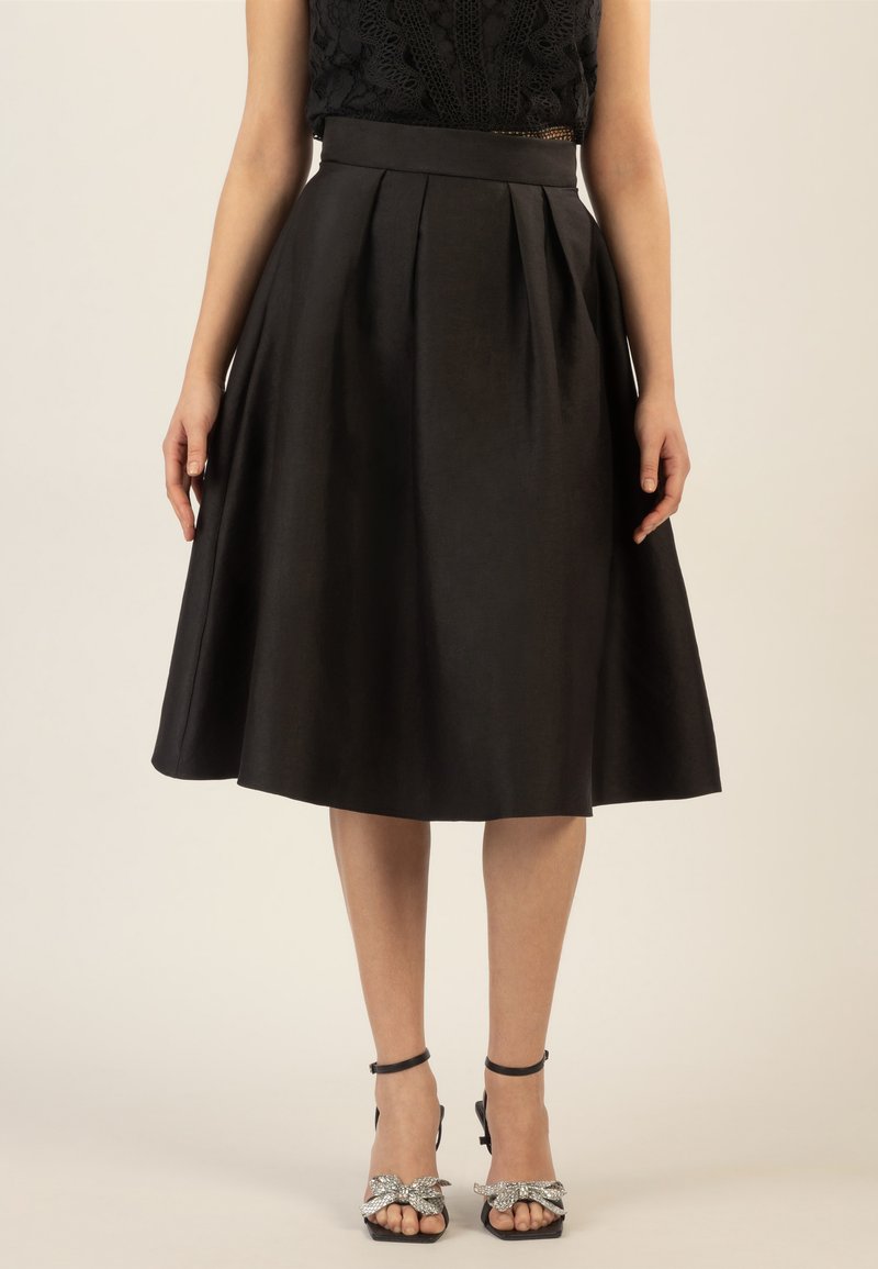 Jupe noire de longueur genou avec des plis, fabriquée en un matériau lisse. Présente une taille ajustée et une silhouette fluide. Associée à des talons noirs à lanières.