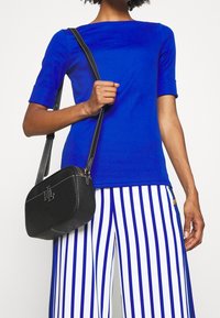 Svart lädercrossbodyväska med vitt sydda sömmar, rektangulär form, med ett präglat logotyp. Kombinerad med en blå topp och randiga byxor.