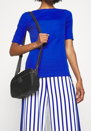 Černá kožená crossbody taška s bílým šitím, obdélníkového tvaru, s vyraženým logem. Kombinováno s modrým topem a pruhovanými kalhotami.