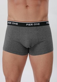 Mand iført tætsiddende mørkegrå boxershorts med sort linning med teksten "PIER ONE" på en lys baggrund.
