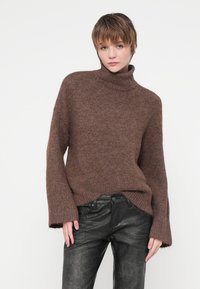 Pull en maille marron à col roulé surdimensionné, avec une texture douce et des poignets côtelés, associé à un pantalon en cuir noir taille haute.
