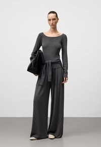 Maglietta grigia a maniche lunghe con scollatura ampia, abbinata a pantaloni grigi larghi. La modella tiene in mano una borsa nera, su uno sfondo neutro.
