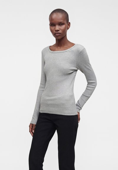 Femme aux cheveux courts portant un haut moulant à manches longues gris clair et un pantalon noir, debout devant un fond blanc uni.