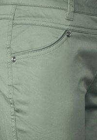 Zelené látkové kalhoty s detailním záběrem na přední část kapsy, s kovovými knoflíky a kontrastním prošíváním.