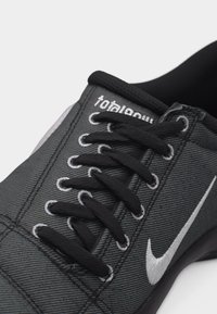 Κοντινό πλάνο μαύρου και γκρι αθλητικού παπουτσιού Nike με μαύρα κορδόνια, λευκό λογότυπο Swoosh και κείμενο "ολική αγωνία" στη γλώσσα.