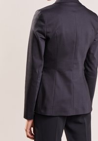 Blazer sartoriale blu con spalle strutturate, lucentezza sottile e linee pulite. Presenta pinces posteriori per una vestibilità ottimale; abbinato a pantaloni scuri coordinati.