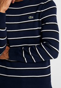 Tmavě modrý svetr s vodorovnými bílými a černými pruhy, žebrovaným kulatým výstřihem a manžetami, s malým zeleným krokodýlím logem na hrudi.