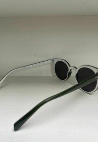 Occhiali da sole rotondi con montatura trasparente e lenti scure. Astine verdi che si assottigliano verso le punte eleganti. Design minimalista con dettagli bicolore.