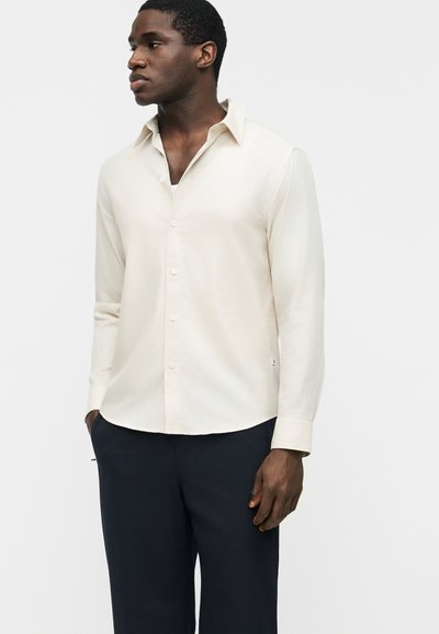 Άνδρας που φοράει ένα μακρυμάνικο κρεμ πουκάμισο με γιακά και κουμπιά, συνδυασμένο με σκούρο παντελόνι, όρθιος με τα χέρια στις τσέπες, κοιτάζοντας πλάγια.