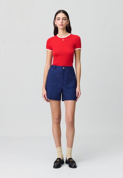 Top corto de rayas rojas con mangas cortas y ribete blanco, combinado con pantalones cortos azul marino que tienen dos bolsillos frontales y acentos dorados. Mocasines negros con calcetines.
