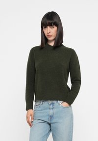 Mørkegrønn strikkegenser med høy hals og lange ermer, med en avslappet passform og myk tekstur, kombinert med lyseblå jeans.
