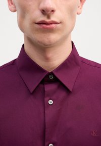 Burgunderfarget skjorte med spredt krage, utstyrt med fem knapper, glatt tekstur og en liten logo på venstre side.