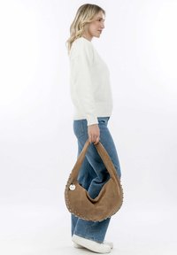 Brun ruskind hobo-taske med en afslappet design, sølvnitter som accentuerer kanten, og en rund form. Båret af en person i en hvid sweater og jeans.