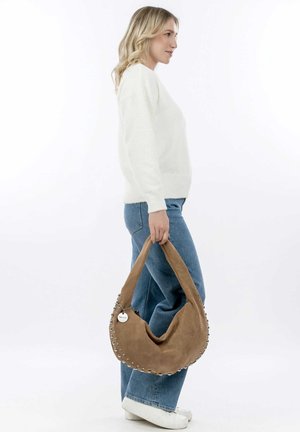 Brown suede hobo bag with a slouchy design, silver studs accenting the edge, and a rounded shape. Worn by a person in a white sweater and jeans.