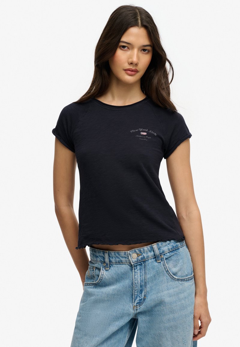 Mujer con una camiseta negra de manga corta con el texto "New York State" y jeans de tiro alto azul claro, de pie contra un fondo blanco.