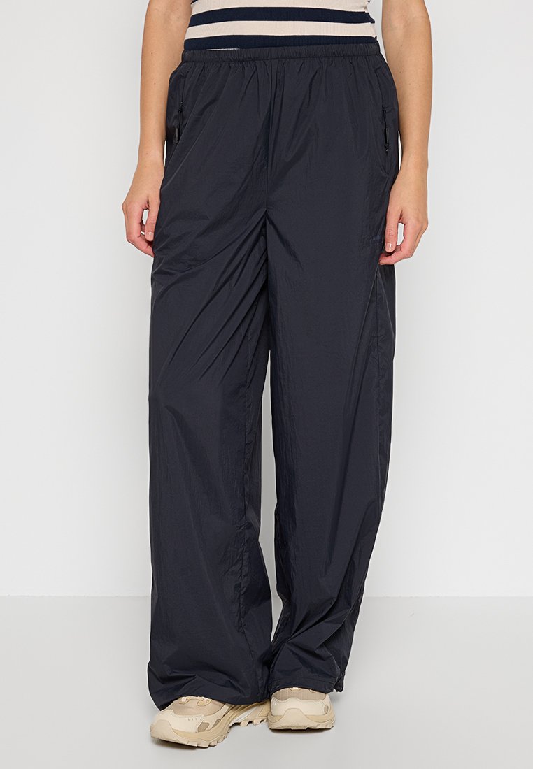 Vrouw draagt losse zwarte nylon broek met elastische tailleband en met rits voorziene zakken, gecombineerd met beige chunky sneakers.