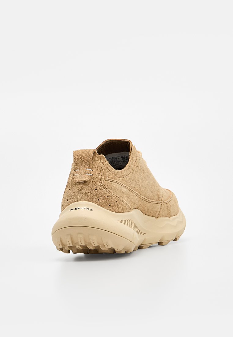 Zapatilla deportiva beige de ante sin cordones con suela gruesa y texturizada y lengüeta en el talón, inclinada para mostrar vista trasera y lateral.