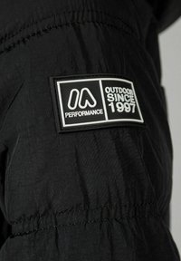 Melnas āra jakas audums ar teksturētu apdari. Iespējas: taisnstūra formas logotipa uzlīme baltā krāsā, uz kuras ir rakstīts "Performance" un "Outdoor since 1997."