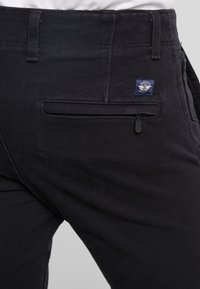 Pantalones de algodón negros con un bolsillo trasero con cremallera, detalles cosidos y una pequeña etiqueta de logo azul en la cinturilla.