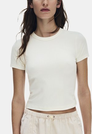 Kvinde iført en tætsiddende, kortærmet, ribbet hvid cropped T-shirt kombineret med lyse beige snorebukser mod en ensfarvet baggrund.