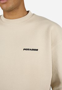 Beige textured sweatshirt with a ribbed collar and black "PEGADOR" logo on the front. The fabric has a subtle diagonal pattern.