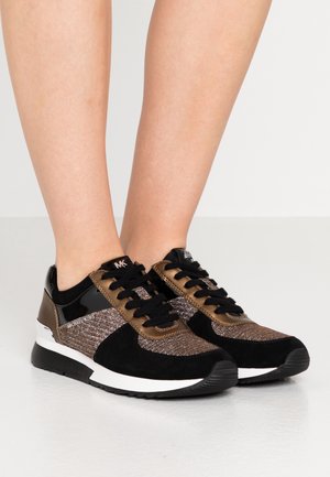 Schwarze und bronzefarbene glitzernde Sneaker mit schwarzen Schnürsenkeln, getragen an nackten Füßen, von der Seite und von vorne auf einer weißen Oberfläche gezeigt.