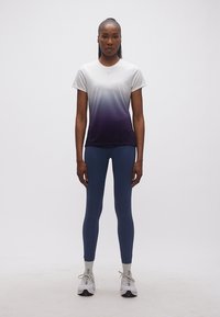 Γυναίκα όρθια, στραμμένη προς τα εμπρός, φορούσα κοντομάνικο πουκάμισο με διαβάθμιση από λευκό σε σκούρο μοβ, ναυτικό μπλε κολάν, λευκές κάλτσες και αθλητικά παπούτσια.