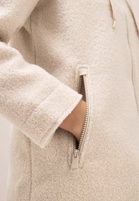 Mano nella tasca di una giacca in pile di colore crema con zip visibile e dettagli sui polsini.