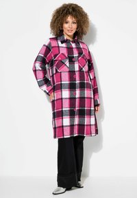 Růžovo-černý károvaný fleece kabát s zapínáním na knoflíky, dvěma náprsními kapsami a dlouhými rukávy; v kombinaci s černými kalhotami a stříbrnými botami.