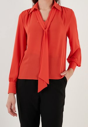 Femme portant un chemisier orange vif à manches longues avec un détail noué au col et un pantalon noir, une main dans la poche, sur un fond uni.