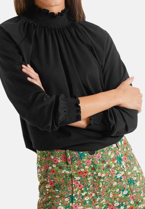Femme portant un haut noir à manches longues avec un col et des poignets à volants, associé à un pantalon vert taille haute avec un motif floral coloré.