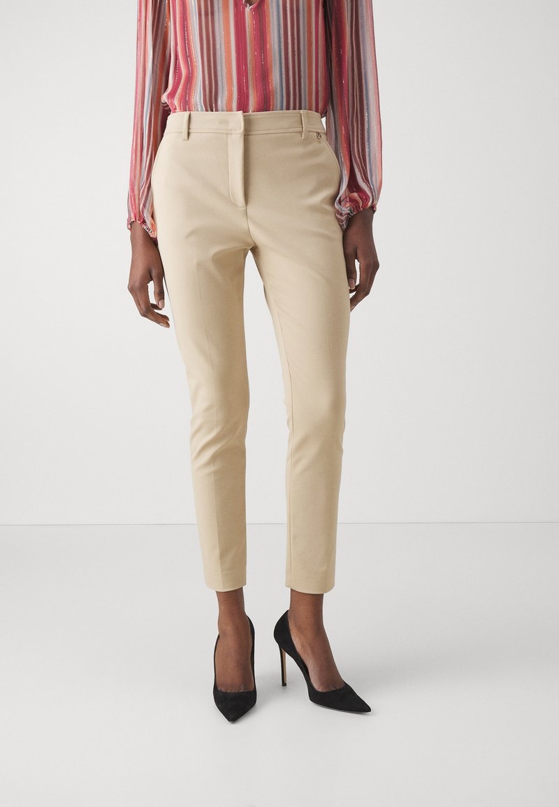 Pantalon beige fuselé à fini lisse, avec plis à l'avant et passants pour ceinture, associé à des escarpins noirs à bouts pointus et un chemisier rayé multicolore.
