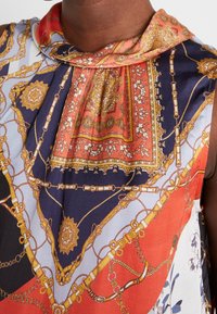 Zijden blouse met een mix van rode, marineblauwe en gouden patronen, versierd met kettingmotieven en bloemaccenten. Zachte textuur, gedrapeerde halslijn.