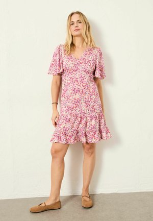 Blumenkleid in Rosa- und Beigetönen, mit kurzen Rüschenärmeln, V-Ausschnitt und einem Volant am Saum. Kombiniert mit braunen flachen Schuhen.