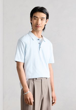 Νέος άνδρας με ανοιχτό μπλε πόλο με κοντά μανίκια και μπεζ πλισέ παντελόνια στέκεται μπροστά σε ανοιχτό γκρι φόντο.