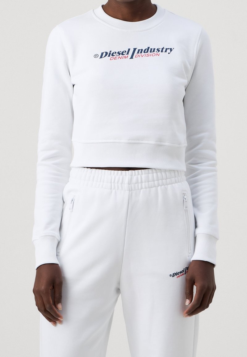 Personne portant un haut court à manches longues blanc et un pantalon de jogging assorti avec les logos "Diesel Industry Denim Division" sur la poitrine et la cuisse.