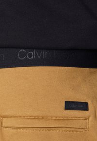 Nahaufnahme einer Senfgelb-Hosentasche mit schwarzem Calvin Klein Etikett und schwarzem Bund mit dezentem Calvin Klein Logo auf einem schwarzen Shirt.