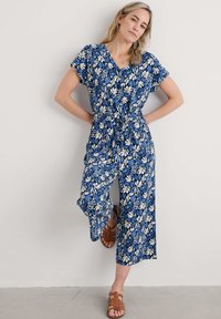 Niebieski kombinezon kwiatowy z dekoltem w kształcie litery V i krótkimi rękawami, zdobiony białymi i jasnoniebieskimi wzorami liści, z wyszczuplającym pasem i szerokimi nogawkami.