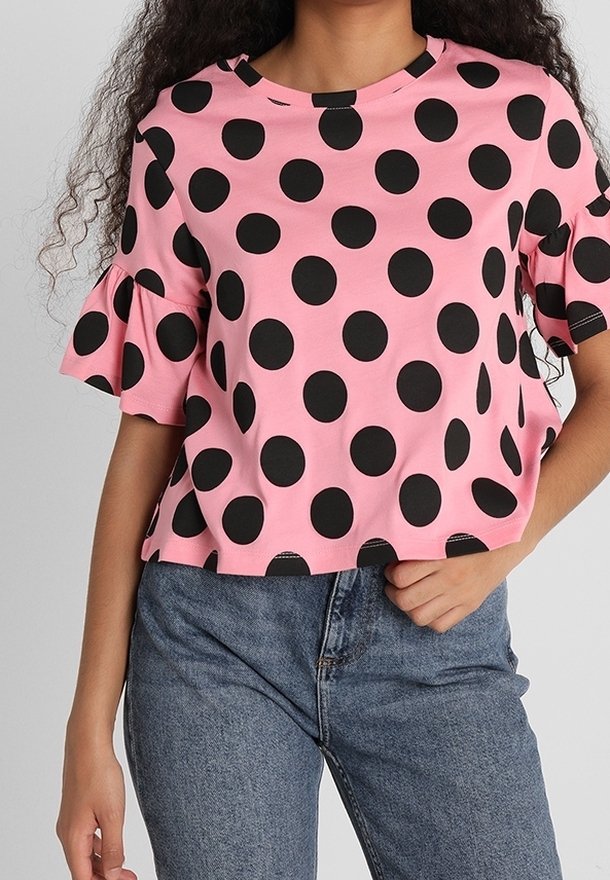 Personne portant un haut rose à gros pois noirs et manches courtes à volants, associé à un jean bleu taille haute.