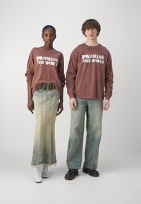 Brune langermede skjorter med "LOFTE VERDEN" grafikk, parret med et grønn-blått denimskjørt og vide jeans. Hvite ankelboots.
