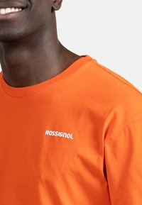 Orange bomuld T-shirt med rund hals og korte ærmer. Har et hvidt "ROSSIGNOL" logo på venstre brystområde. Glat tekstur.
