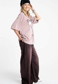 Oversizowa różowa koszulka z nadrukiem na krótki rękaw, zestawiona z szerokimi fioletowymi spodniami. Modelka nosi czarną czapkę i stoi bokiem.