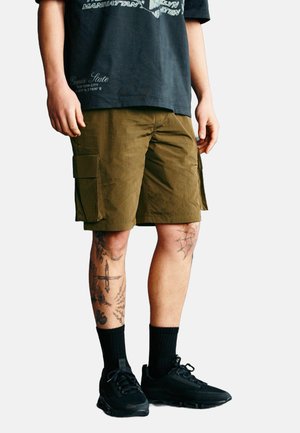 Khaki cargo šortky s více bočními kapsami, doplněné volným černým tričkem s grafikou a černými teniskami, které mají krátké černé ponožky.