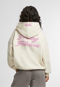 Felpa cremosa con texture cosparsa, che presenta la scritta rosa "Feeling Delicious" sul retro e un dettaglio a fiocco sul cappuccio.